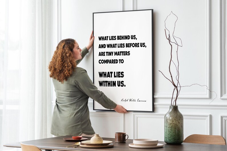 Puede incluir: Una impresi&oacute;n enmarcada con la cita "WHAT LIES BEHIND US, AND WHAT LIES BEFORE US, ARE TINY MATTERS COMPARED TO WHAT LIES WITHIN US." Una mujer cuelga el arte enmarcado en negro en una pared blanca. Un jarr&oacute;n y una mesa est&aacute;n en primer plano.