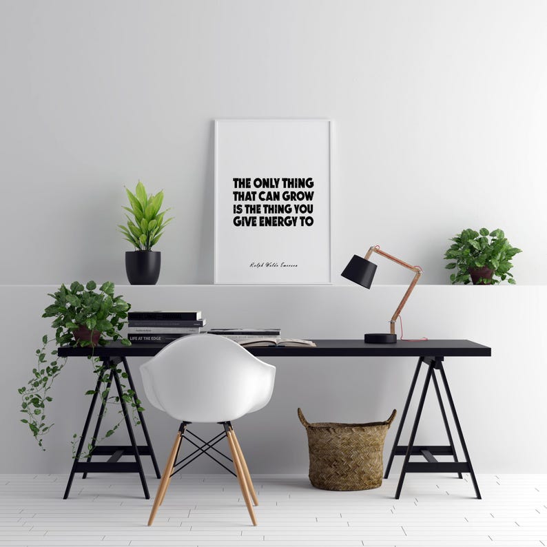 May include: A modern home office setup featuring a black desk with a white chair and a framed quote. The desk is supported by black trestle legs and includes a copper and black desk lamp, a woven basket, and potted plants.