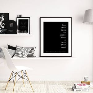 May include: Black and white typography print with the quote "You have been in every line I have ever read" by Charles Dickens. The print is framed and hanging on a white wall in a modern living room.