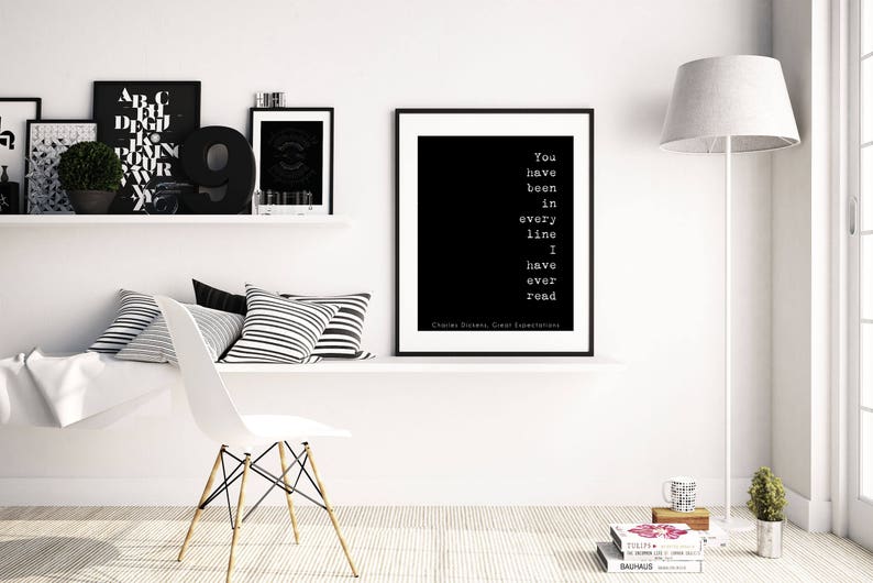 May include: Black and white typography print with the quote "You have been in every line I have ever read" by Charles Dickens. The print is framed and hanging on a white wall in a modern living room.