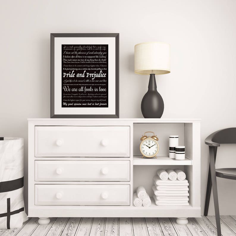 May include: A black and white framed print with a white background and black text. The text is a quote from the novel "Pride and Prejudice" by Jane Austen. The print is sitting on a white dresser with a black lamp and a gold alarm clock.