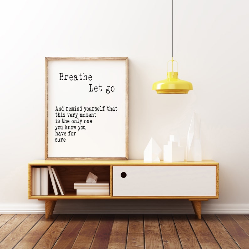 May include: A framed print with a white background and black text that reads "Breathe Let go And remind yourself that this very moment is the only one you know you have for sure". The print is hanging on a white wall above a wooden shelf with a white drawer. There are white decorative objects on the shelf.