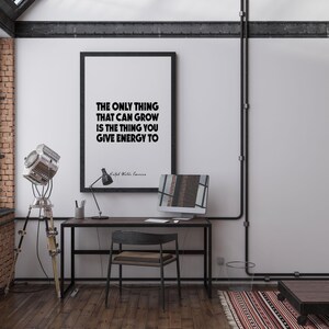 May include: An interior shot of a modern office space with a framed print that reads "THE ONLY THING THAT CAN GROW IS THE THING YOU GIVE ENERGY TO." The room features a desk, chair, and a vintage-style spotlight on a tripod.