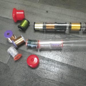 May include: Assortment of colorful thread spools in a clear plastic container. The spools are arranged in a row and include gold, silver, red, and green thread.