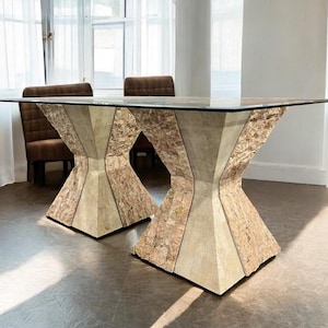 May include: A contemporary dining table with a clear glass top. The base has two geometric supports made of light brown and beige textured materials. The table is in a bright room with sheer curtains and two brown upholstered chairs.