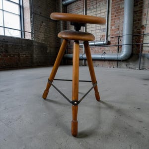 May include: A wooden stool with a round seat and a circular backrest. The stool features three tapered legs connected by a black metal triangular support frame. The seat height is adjustable via a central black metal screw mechanism.