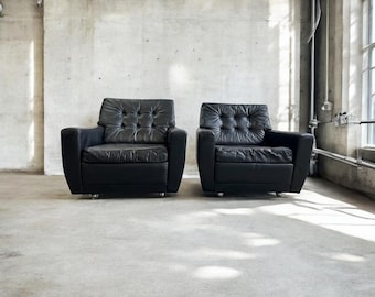 May include: Two black leather armchairs with button tufting and a mid-century modern design. The chairs are positioned in front of a concrete wall.