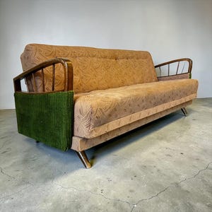 Puede incluir: Un sofá vintage con tela beige estampada y reposabrazos de madera. El sofá presenta un panel de pana verde en el lateral y patas de madera en ángulo. Los cojines del respaldo y del asiento tienen un diseño de remolino repetido.