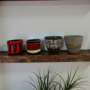 Op de afbeelding: Vier keramische potten met unieke ontwerpen staan op een donkere houten plank. De potten hebben verschillende kleuren, waaronder rood, bruin en grijs, met geometrische en abstracte patronen. De potten zijn van verschillende maten en vormen.
