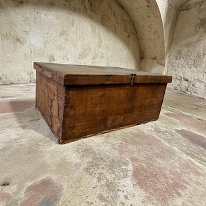 Könnte beinhalten: Eine rechteckige Holzkiste mit Scharnierdeckel. Die Kiste besteht aus dunkelbraunem Holz mit sichtbarer Maserung und einem Metallverschluss. Die Kiste wirkt gealtert, mit einer rustikalen Ästhetik. Die Kiste wird wahrscheinlich zur Aufbewahrung verwendet.