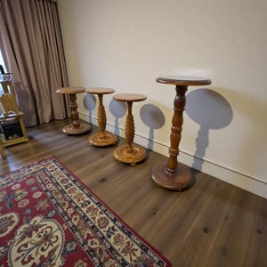 Könnte beinhalten: Vier Pflanzenständer aus Holz mit runden Oberflächen und verzierten, gedrechselten Beinen. Die Ständer haben unterschiedliche Designs, einige mit einem gedrehten, seilartigen Muster auf der Mittelsäule. Die Ständer sind aus warm getöntem Holz gefertigt.