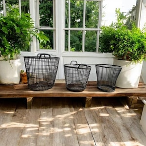 Puede incluir: Tres cestas de alambre negro de diferentes tamaños con asas están sobre una superficie de madera. Las cestas tienen un diseño abierto. Plantas verdes en macetas blancas están en el fondo, cerca de una ventana.