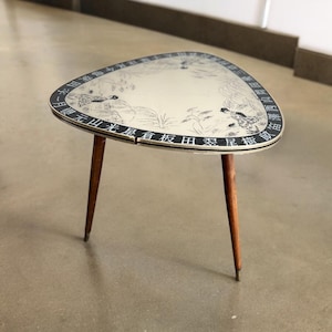Puede incluir: Una pequeña mesa triangular de estilo moderno de mediados de siglo con una tapa blanca y un diseño pintado en negro. La mesa tiene tres patas de madera y presenta un diseño en blanco y negro con caracteres chinos alrededor del borde.