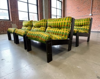 May include: A set of four mid-century modern armchairs. The chairs have dark wood frames and feature cushions with a yellow, green, and brown plaid pattern. The chairs are arranged in a row against a brick wall.