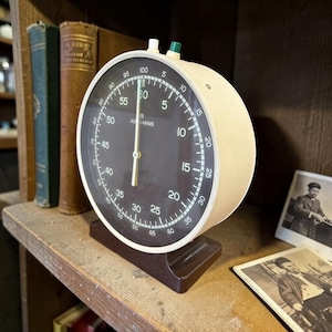 May include: A vintage stopwatch with a black face and white numerals, marked with the brand "JUNGHANS". The stopwatch has a cream-colored casing and sits on a brown base. The time is set to 60 seconds. The stopwatch is on a wooden shelf.