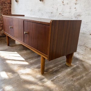 Puede incluir: Un aparador de madera de estilo moderno de mediados de siglo con dos puertas y tiradores de latón. El mueble es de color marrón oscuro y se apoya en cuatro patas cónicas. El diseño es sencillo y elegante, con líneas limpias y una estética minimalista.