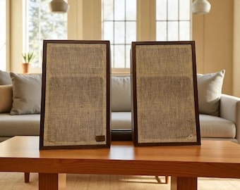 May include: Two vintage-style speakers with dark brown wooden frames and tan fabric fronts sit on a wooden coffee table. The speakers are rectangular and have a classic, retro design, perfect for home audio.