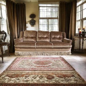 May include: A brown velvet sofa with fringe trim sits in a room with a patterned rug, brown curtains, and a window with a view of trees.