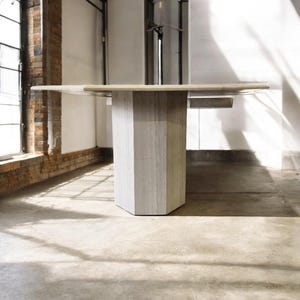 Op de afbeelding: Een moderne stenen tafel met een geometrische sokkel. Het rechthoekige tafelblad heeft een lichtbeige kleur. De basis is een lichtgrijze, veelzijdige kolom. De tafel is ongeveer 180 cm lang.