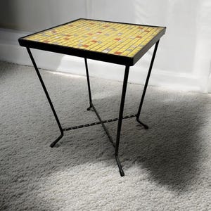 May include: A small, square side table with a mosaic tile top in yellow, red, and gray. The table has a black metal frame and legs. The table is approximately 18 inches tall.