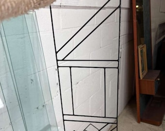 May include: Black metal magazine rack with a geometric design. The rack is made of thin metal rods that are welded together to create a series of rectangular and triangular shapes.