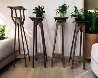 May include: Four antique wooden plant stands of varying heights and designs. Each stand has a dark brown finish and a small shelf. The stands hold potted green plants, creating a natural, rustic aesthetic.