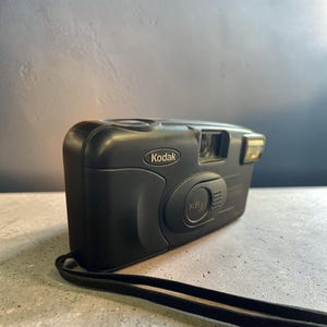 Peut inclure: Un appareil photo argentique Kodak KB10 noir au format 35 mm, doté d'un boîtier rectangulaire compact, d'un objectif central, d'un flash intégré sur la droite, d'un viseur et d'une dragonne noire fixée au coin inférieur de l'appareil.