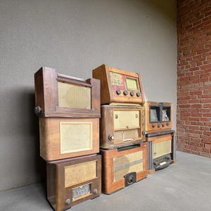 Op de afbeelding: Een verzameling vintage houten radio's die op elkaar zijn gestapeld. De radio's hebben verschillende ontwerpen, waaronder luidsprekerroosters, wijzerplaten en afstemindicatoren. De houttinten variëren van licht tot donker, sommige radio's vertonen tekenen van ouderdom.
