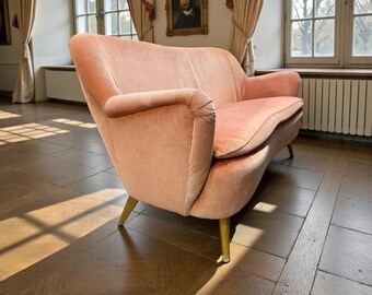 Puede incluir: Un sofá vintage de terciopelo color rubor con respaldo y reposabrazos curvos. El sofá tiene tres patas doradas y está situado en una habitación con grandes ventanales y suelo de madera. Es un mueble.