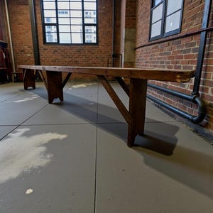 Op de afbeelding: Een lange, donkerbruine houten bank. De bank heeft een eenvoudig ontwerp met schuine poten en een rechthoekige zitting. Hij staat voor een bakstenen muur en een raam. De bank is ongeveer 200 cm lang.