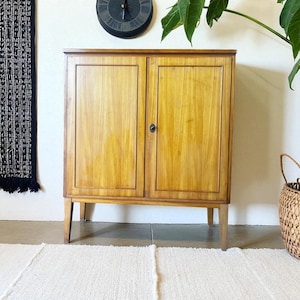 Puede incluir: Un mueble de madera clara con dos puertas y un pequeño tirador redondo. El mueble tiene un diseño sencillo con cuatro patas finas. Un reloj negro cuelga de la pared encima del mueble.