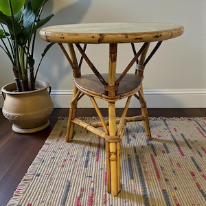 May include: A round bamboo side table with a light-colored top and a lower shelf. The table has a natural bamboo finish and is supported by four legs. The table is placed on a rug with colorful stripes.