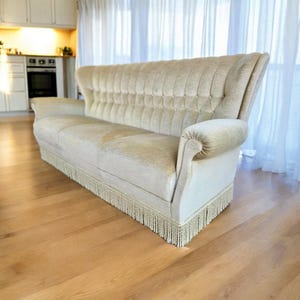 May include: A cream-colored, vintage-style sofa with a high back and rolled arms. The sofa features a tufted backrest and a fringe along the base. The sofa is placed on a hardwood floor.