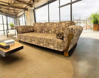 May include: A vintage floral-patterned sofa with wooden accents and a matching bolster pillow. The sofa is set in a room with large windows and a neutral-toned rug. The sofa's fabric features a mix of brown, beige, and green floral designs.