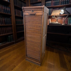 Könnte beinhalten: Ein hoher, antiker Aktenschrank aus Holz mit Rollladentür. Der Schrank ist aus warm getöntem Holz gefertigt und hat eine kleine Schublade oben. Er steht in einer Bibliothek, umgeben von Bücherregalen.