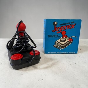 Może przedstawiać: Czarny joystick z czerwonymi przyciskami i czerwoną gałką. Joystick znajduje się obok niebieskiego pudełka z napisem "Joystick" w kolorze czerwonym i tekstem "Competition Pro". Pudełko zawiera również ilustrację joysticka.
