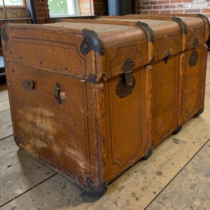 May include: A vintage brown trunk with metal detailing. The trunk is rectangular with a curved top and metal corner protectors. It has a hinged lid and decorative trim. The trunk is likely made of wood and shows signs of age and wear.