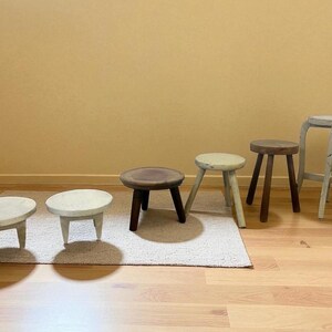 May include: A collection of small wooden stools in various styles and colors. The stools range from light-colored to dark brown, with different leg designs and seat shapes. Some are painted, others are natural wood.