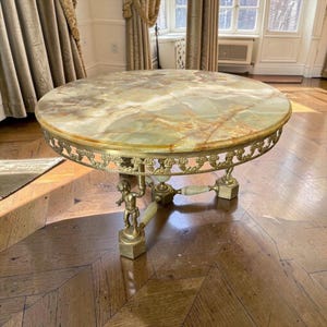 May include: A round coffee table featuring a polished green and brown onyx stone top. The table has a decorative gold-toned metal filigree apron and three legs, each designed with a small metal cherub figure and onyx accents on a base.