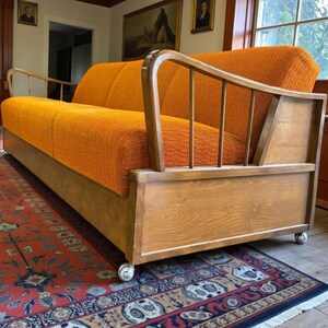 May include: An orange upholstered sofa with a wooden frame and armrests. The sofa sits on a wooden base with small wheels. The sofa is placed on a patterned rug.
