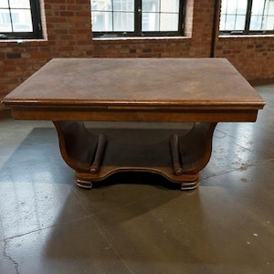 Puede incluir: Una mesa de centro cuadrada de madera grande con un acabado marrón oscuro. La mesa tiene un diseño de base curvada único y se asienta sobre un suelo de hormigón. La mesa está en una habitación con paredes de ladrillo y grandes ventanas.