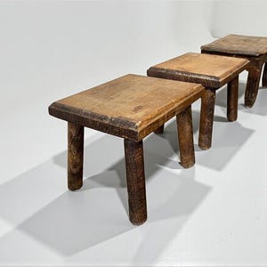 May include: Three small, rustic wooden stools with rectangular seats and rounded legs are arranged in a row. The stools are made of aged wood, displaying a natural, weathered appearance. The stools are approximately 10 inches long, 6 inches wide, and 8 inches tall.