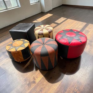 Puede incluir: Una colección de cinco otomanas de cuero que incluye un cubo negro cuadrado, una otomana circular beige y negra con patrones de estrellas, una otomana circular a rayas beige y marrón, una otomana con patrón triangular marrón y negro, y una otomana roja y negra con estrellas.