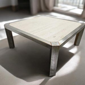 Peut inclure: Table basse carrée avec un dessus clair et texturé et un cadre en métal brossé. La table a quatre pieds robustes et un design simple et moderne. La table mesure environ 91 cm de côté.