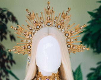 Könnte beinhalten: Eine goldene Krone mit einem Sternenausbruch-Design und ein goldenes Ketten-Körpergeschirr mit blauen und weißen Perlen. Die Krone und das Geschirr befinden sich auf einem weißen Mannequin.