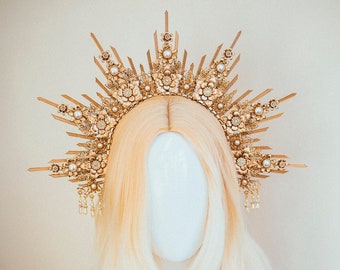 May include: A gold crown with pearl accents and a sunburst design. The crown is on a white mannequin head with long, blonde hair.