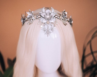Può includere: Una corona argentata con decorazioni floreali e a forma di stella. La corona è realizzata in metallo e ha un design delicato ed etereo.