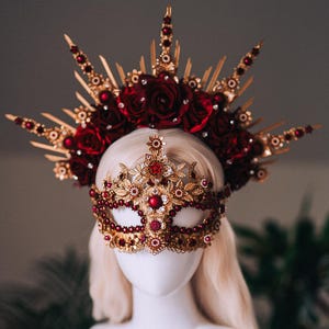 Puede incluir: Un conjunto de tocado y máscara dorados con detalles rojos. El tocado presenta un diseño de rayos de sol con rosas rojas y adornos brillantes. La máscara está adornada con gemas rojas e intrincados filigranas dorados.