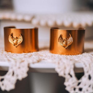 May include: Two copper-colored cuff bracelets with a gold-colored scarab beetle and wing design. The bracelets are displayed on a white surface with a lace doily. The bracelets are a unique accessory.