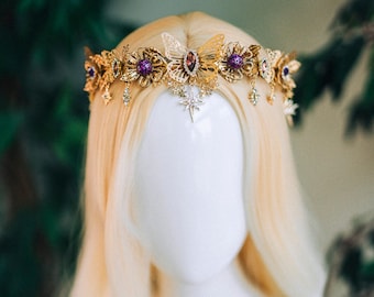 Puede incluir: Una corona dorada con gemas púrpuras y detalles de mariposa. La corona está sobre una cabeza de maniquí blanca con cabello largo y rubio.
